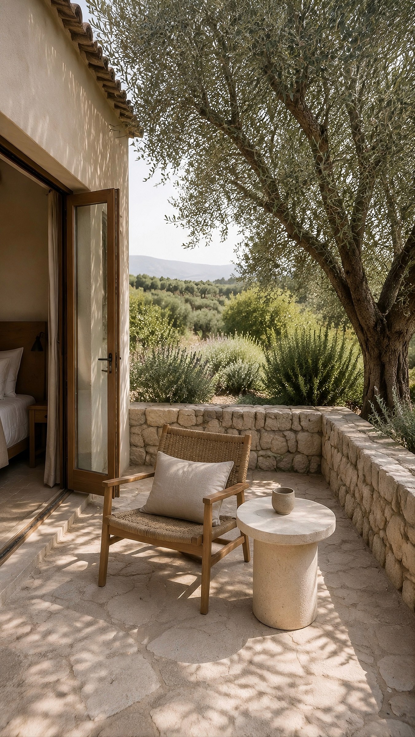 Stone terrace with a woven lounge chair and round side table under the shade of an olive tree.
