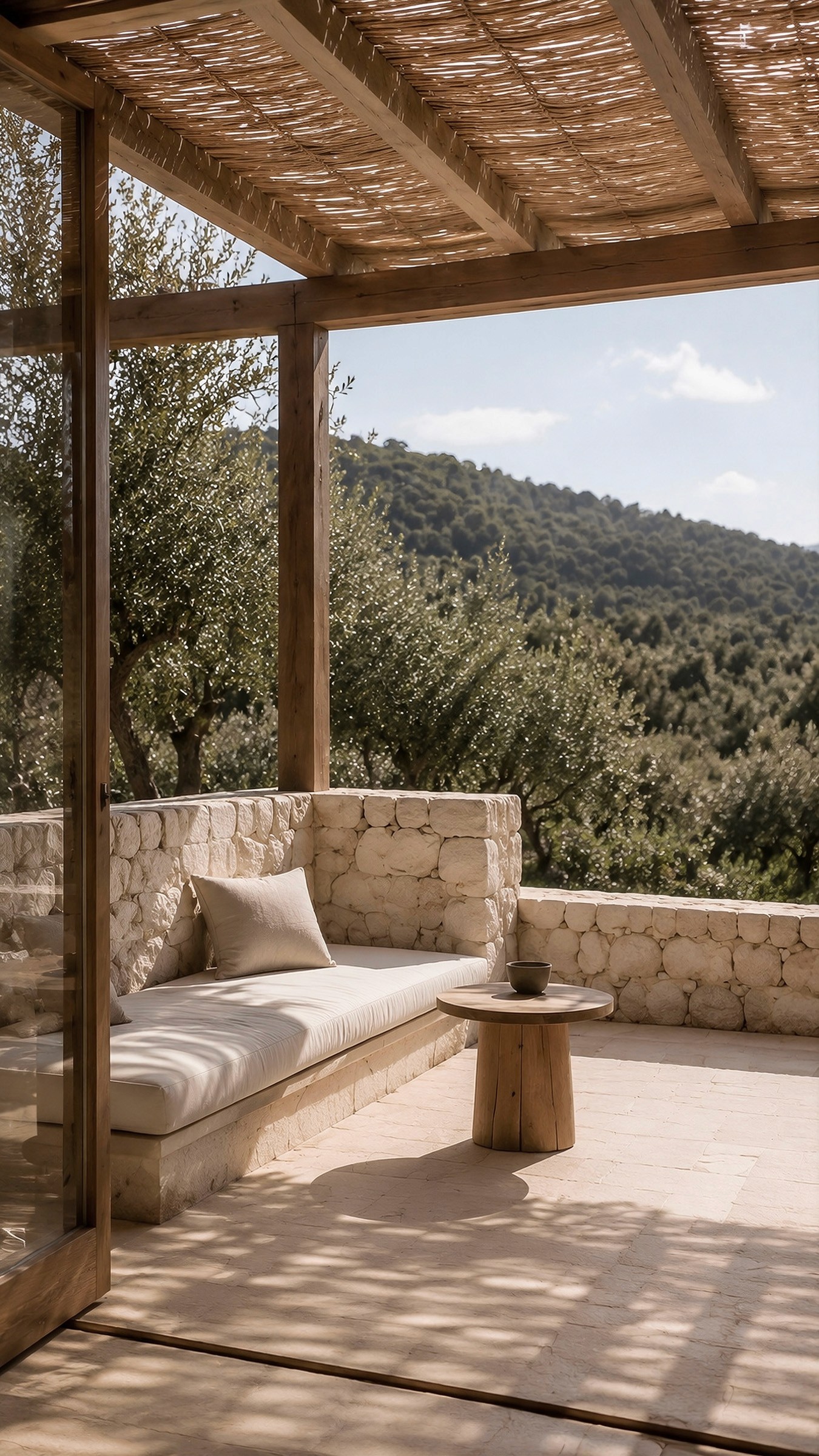 Shaded stone terrace with a built-in stone bench, cushions, and a small wooden table, overlooking a forest.