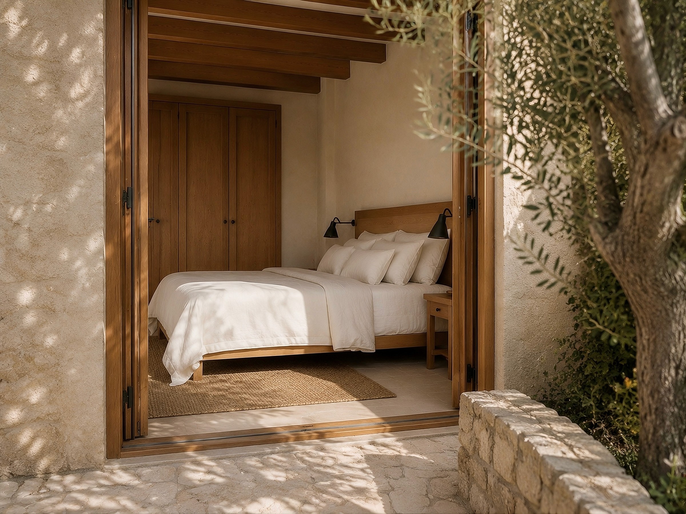 View into a bright bedroom with a wooden bed, seen through open folding doors from a stone patio.